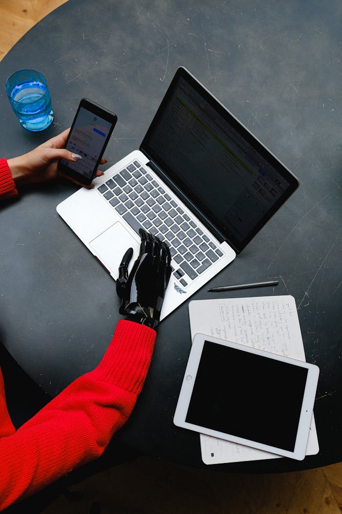 who-we-are A prosthetic hand uses a laptop on a workspace with gadgets and notes around.